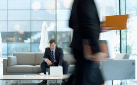 Businessman sitting in modern office lounge using a laptop while colleague walks past, creating dynamic blur. — 5 Proven Mess