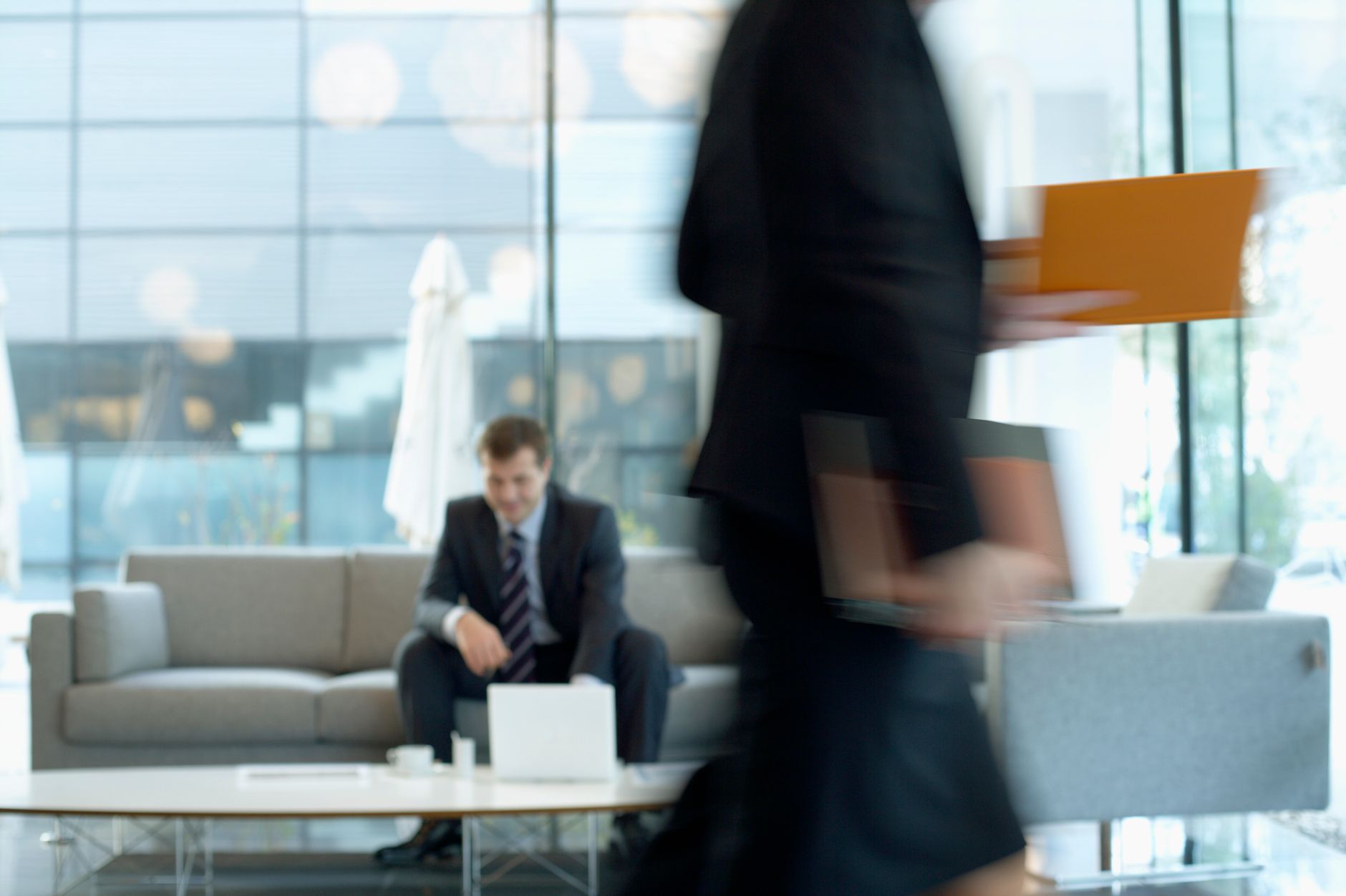 Businessman sitting in modern office lounge using a laptop while colleague walks past, creating dynamic blur. — 5 Proven Mess